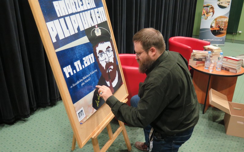 Andrzej Pilipiuk składa autograf na plakacie promującym spotkanie
