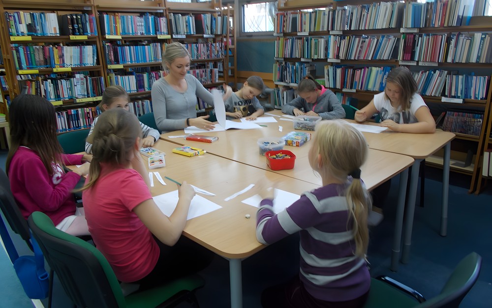 Dzieci wraz z bibliotekarką siedzą przy stole wykonując ilustracje