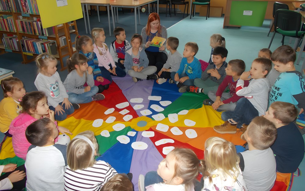 Dzieci siedzą na chuście animacyjnej i słuchają opowiadania czytanego przez bibliotekarkę