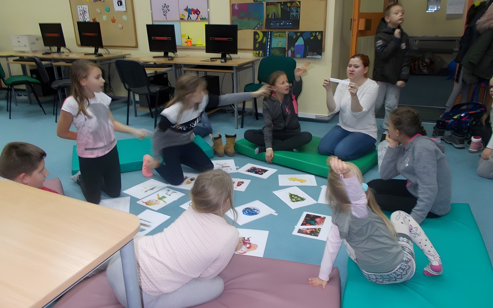 Bibliotekarka pokazuje wyraz po angielsku, dzieci zglaszają się by dopasować wyraz do obrazków leżących na dywanie