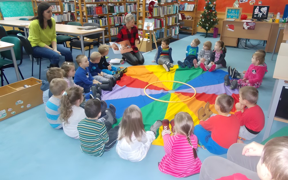 Dzieci siedzą na chuście animacyjnej słuchając książki czytanej przez bibliotekarkę