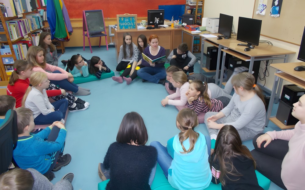 Dzieci siedzą na materacach i słuchają bibliotekarki, która czyta im mit