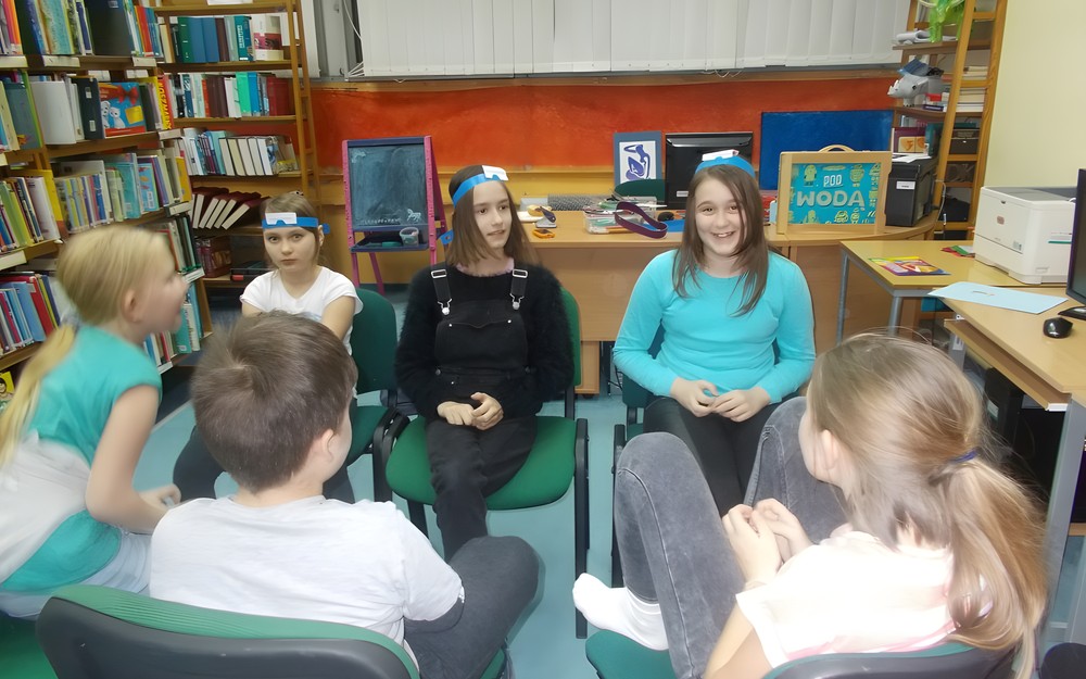 Trzy dziewczynki siedzą na krzesłach z opaskami na głowie i biorą udział w zabawie, natomiast reszta dzieci obserwuje ich poczyniania