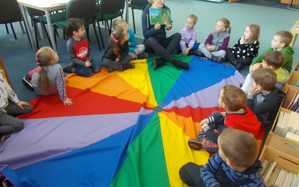 Dzieci siedzą na chuście animacyjnej, a bibliotekarka pokazuje im ilustracje w czytanej książce