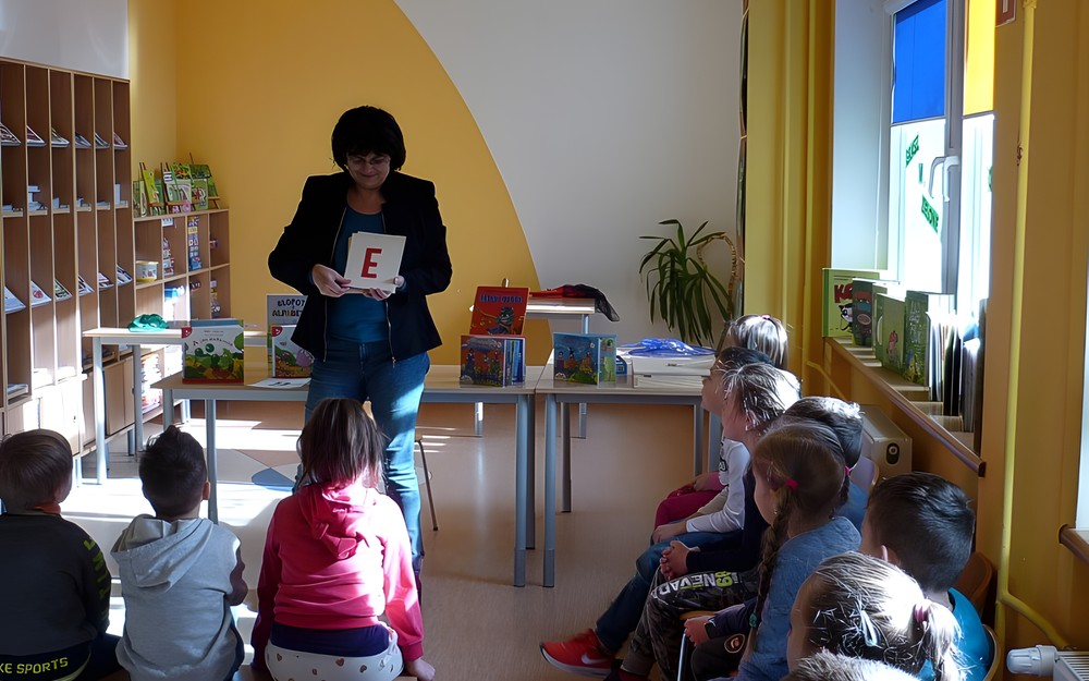 Dzieci siedzą na ławeczce i krzesełkach, bibliotekarka pokazuje literki alfabetu