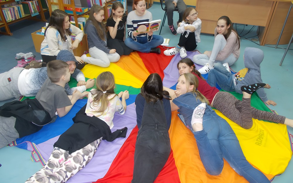 Dzieci  siedzą na chuście animacyjnej i słuchają bibliotekarki czytającej opowiadanie