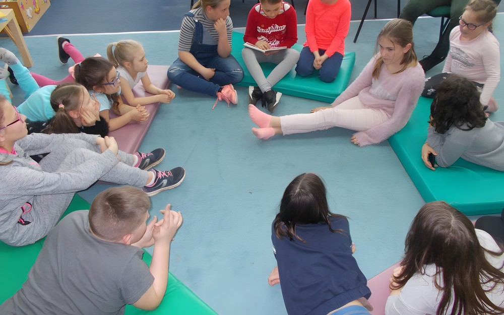 Dzieci siedzą na materacykach i słuchają opowiadania czytanego przez jedną z uczestniczek