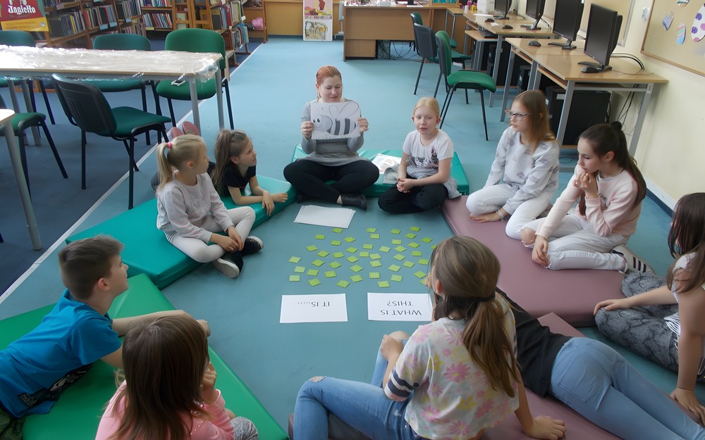 Dzieci siedzą na materacach i odpowiadają na pytanie bibliotekarki w jęzuku angielskim