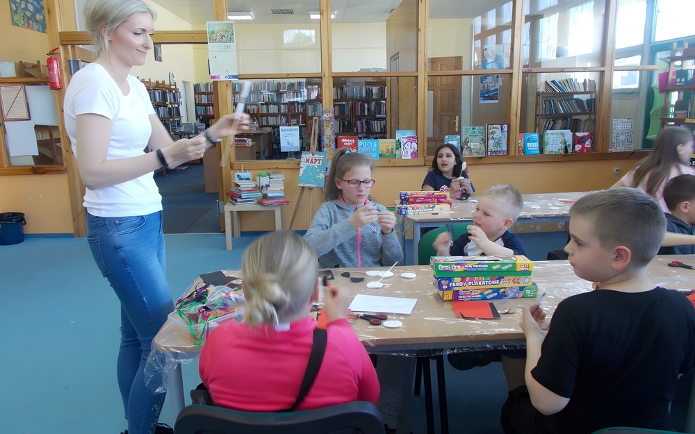 Dzieci siedza przy stole, a bibliotekarka pokazuje jak wykonać pracę plastyczną