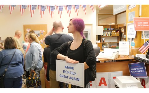 W centrum zdjęcia widoczna młoda kobieta w rózowych włosach z tabliczką Make books great again!