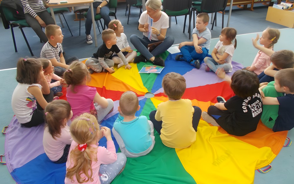 Dzieci siedza na chuście animacyjnej i słuchają czytanego przez bibliotekarkę opowiadania