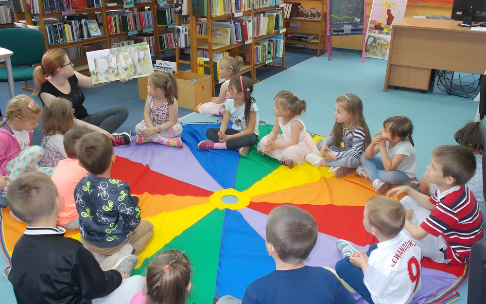 Dzieci siedzą na chuście i słuchają opowiadania czytanego przez bibliotekarkę