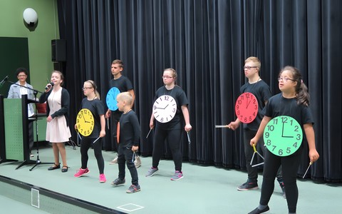 Występ taneczny uczestników cyklu zajęć terapeutycznych w KBP w roku szkolnym 2017/2018. Młodzież i dzieci stoją w dwóch rzędach, mają na szyjach kolorowe wizerunki zegarów, w rękach trzymają trójkąty, ubrani są na czarno, wokalistka na jasno.