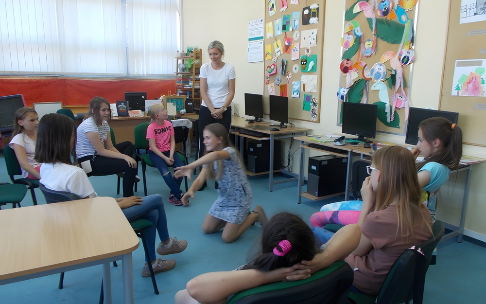 dzieci siedzą na krzesłach obserwując dziewczynkę, która pokazuje pantomimiczne hasło