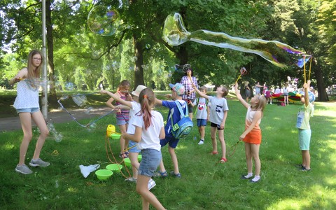Dzieci uczestniczące w akcji Bezpieczne wakacje w bibliotece znajdują się w parku przed budynkiem głównym biblioteki. Dziewczynki i chłopcy robią gigantyczne bańki mydlane
