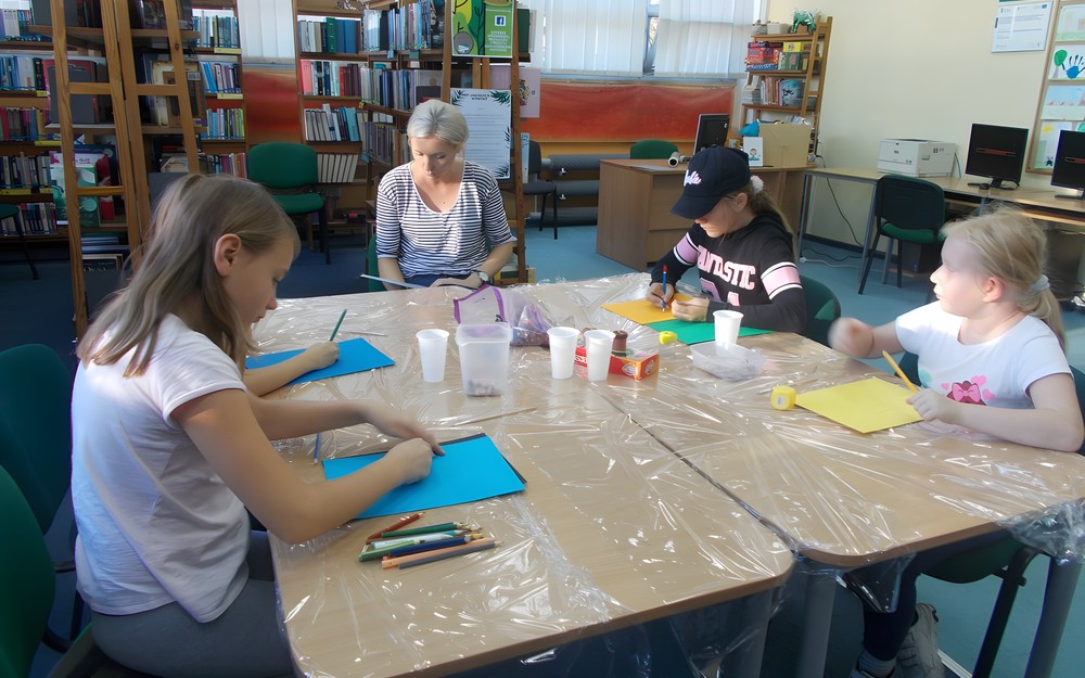 Dzieci siedzą przy stole wykonując prace plastyczne i słuchają czytanego przez bibliotekarkę opowiadania