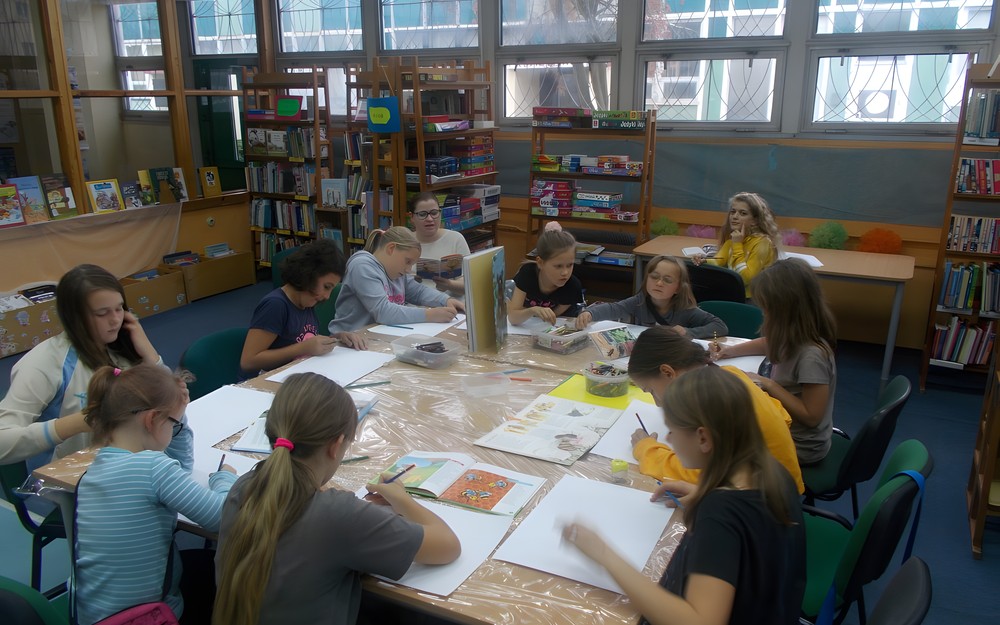 Dzieci siedzą przy stole wykonując prace plastyczne i słuchają czytanego przez bibliotekarkę opowiadania