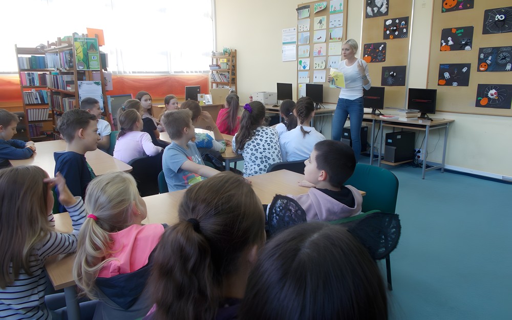 Uczniowie siedzą przy stolikach słuchając opowieści bibliotekarki