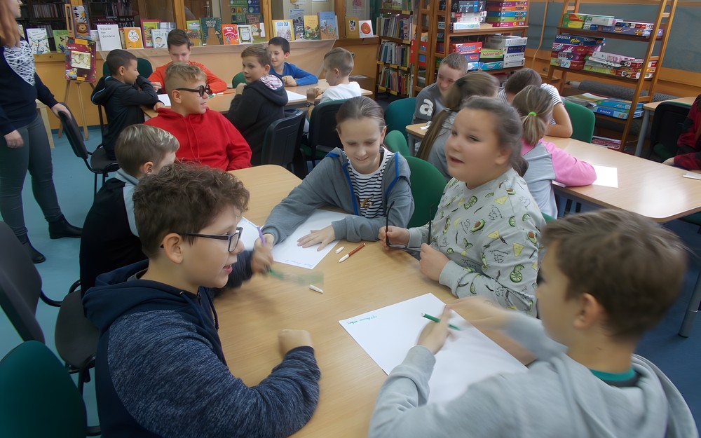 Dzieci siedzą przy stołach i pracują w grupach tworząc reklamy produktów