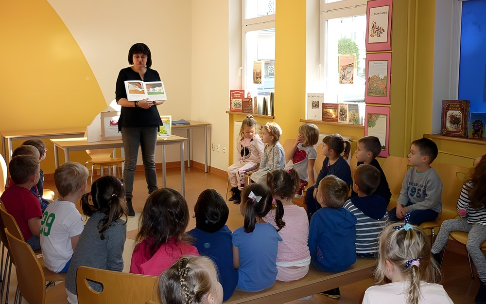 Dzieci siedzą na krzesełkach, bibliotekarka prezentuje książkę Maxa Velthuijs'a JAK DOBRZE BYĆ ŻABKĄ
