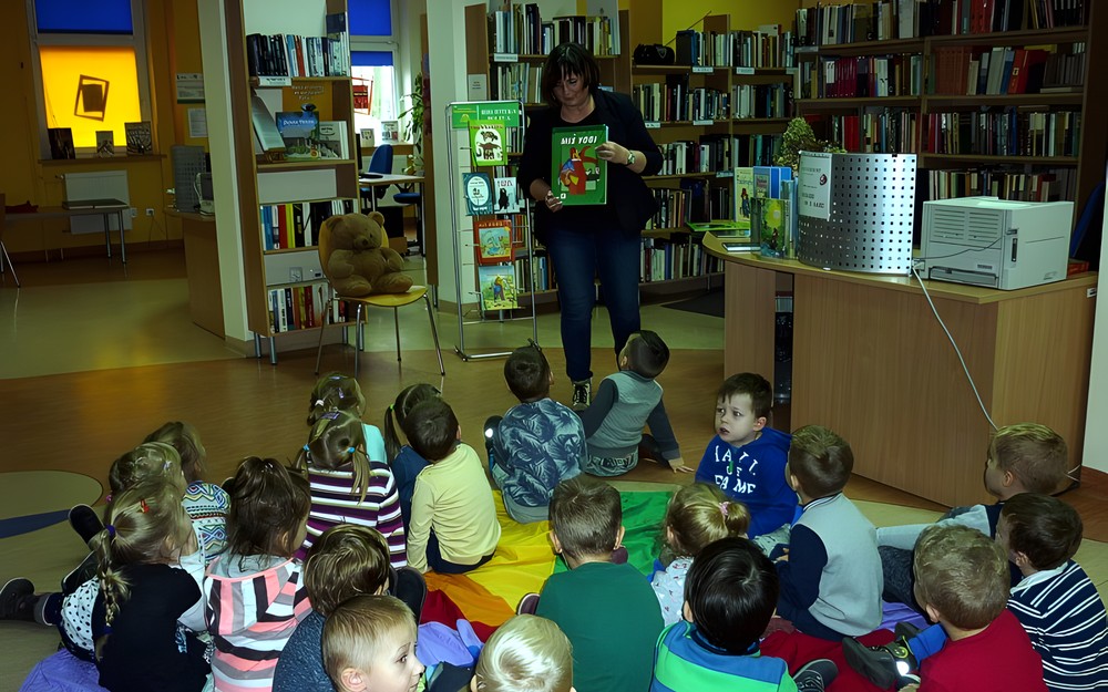 Dzieci siedzą na chuście integracyjnej i przyglądają się książkom o misiach prezentowanych przez bibliotekarkę