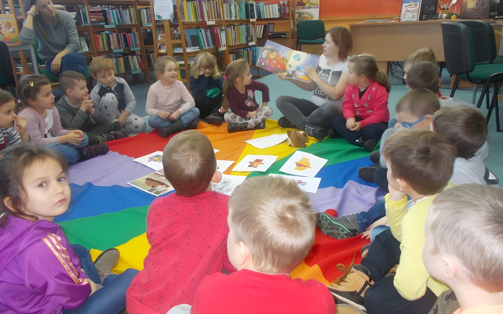 Dzieci siedzą na chuście i słuchają opowiadania czytanego przez bibliotekarza