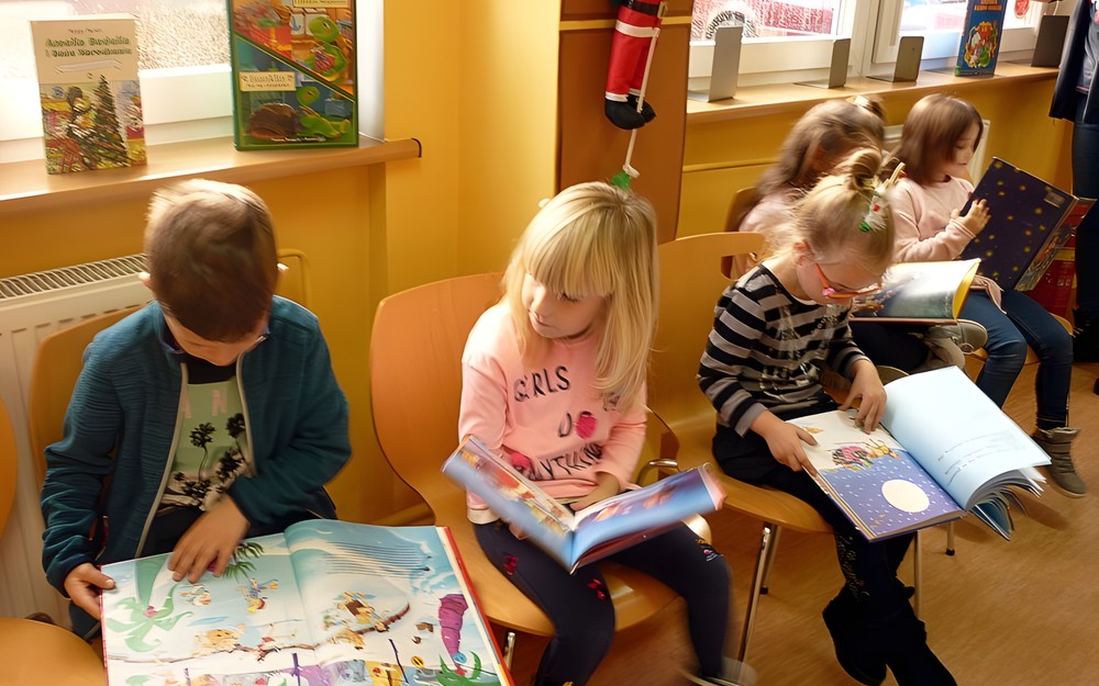 Dzieci siedzą na krzesełkach i przeglądają książki z wystawki BAJKOWE ŚWIĘTA