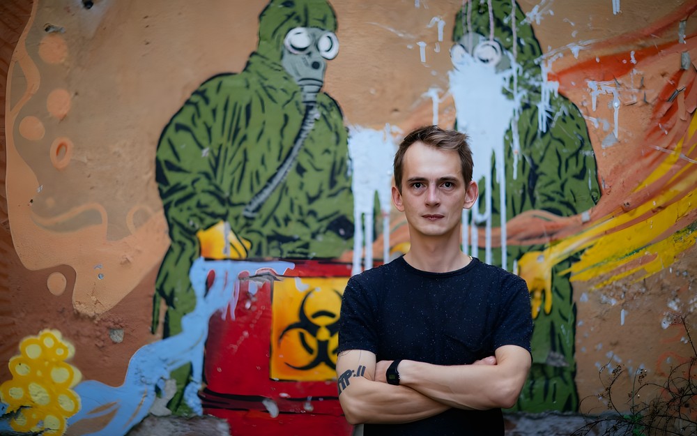 Na zdjęciu: Jasiu Kapela na tle muralu, na którym widnieją dwie postacie w kombinezonach i maskach ochronnych przy skrzyni ze skażonym materiałem. Mural jest oblany błękitną farbą.