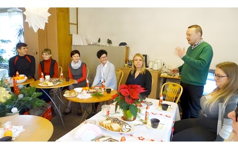 Spotkanie świąteczne czytelników „książek mówionych”. Dyrektor Koszalińskiej Biblioteki Publicznej wita gości zgromadzonych podczas spotkania. Zgromadzeni siędzą przy zastawionych stołach.