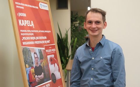Jaś Kapela uśmiechnięty stoi obok tablicy, na której umieszczony jest plakat promujący spotkanie z nim