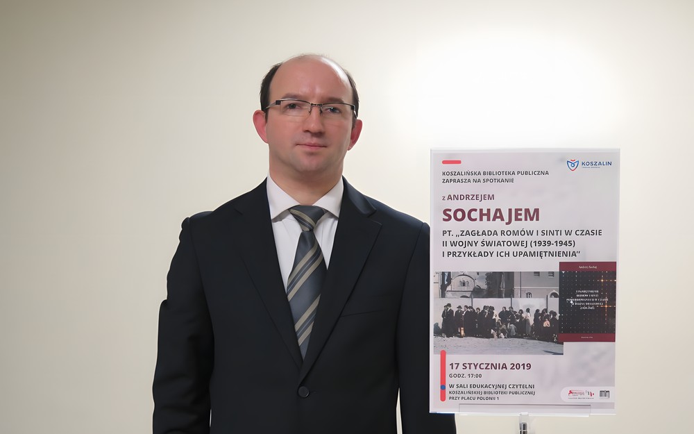 Andrzej Sochaj obok tabliczki z platatem promującym wykład na temat zagłady Romów i Sinti