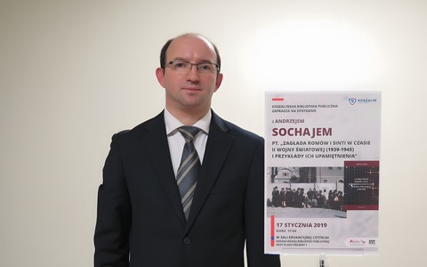 Andrzej Sochaj obok tabliczki z platatem promującym wykład na temat zagłady Romów i Sinti