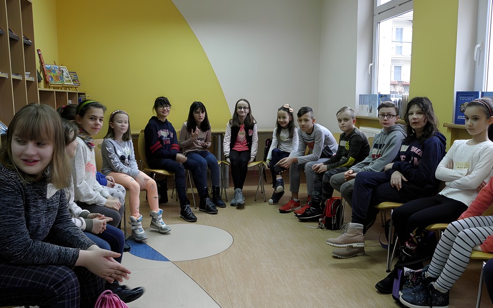 Dzieci siedzą na krzesełkach i czekają na rozpoczęcie zajęć