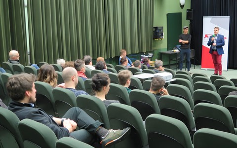 Dwóch mężczyzn wita publiczność zgromadzoną  na projekcji filmu. Na pierwszym planie widać zgromadzoną publiczność.