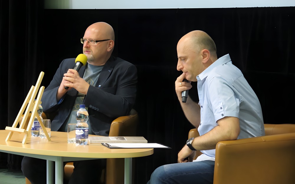 Od lewej: Grzegorz Śliżewski, prowadzący spotkanie  Piotr Polechoński siedzą na brązowych fotelach przy owalnym stoliku.
