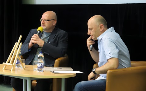 Od lewej: Grzegorz Śliżewski, prowadzący spotkanie  Piotr Polechoński siedzą na brązowych fotelach przy owalnym stoliku.