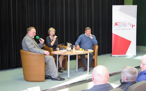 Od lewej: Zdzisław Pacholski, Anna Rawska, Zbigniew Choiński siedzą na fotelach przy stoliku, przed nimi publiczność zgromadzona na spotkaniu.