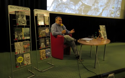 Marcin Kostrzyński siedzi przy stoliku podczas spotkania w Koszalińskiej Bibliotece Publicznej. Z jego prawej strony wyeksponowane są książki o tematyce przyrodniczej. W tle widać prezentację z motywem górskim