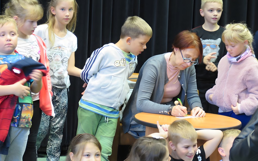 Małgorzata Strękowska-Zaremba otoczona wianuszkiem najmłodszych czytelników siedzi przy okrągłym stoliku i składa autograf.