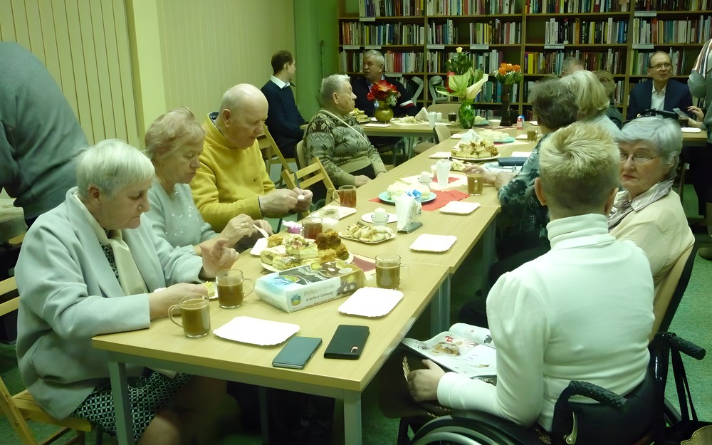 na zdjęciu zebrali siedzą przy stołach i jedzą ciasto