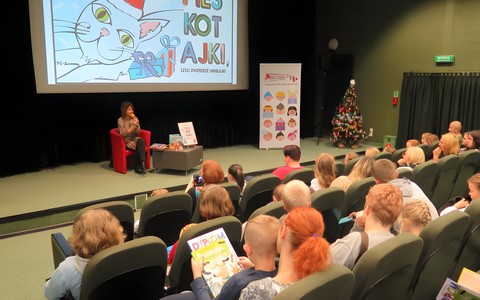 Spotkanie autorskie z Agatą Widzowską. W tle widać autorkę siedzącą w czerwonym fotelu przy stoliku. Na ścianie widać slajd z napisem PiesKotajki. Na pierwszym planie zgromadzona publiczność.