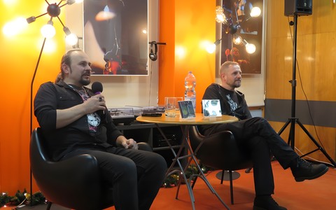 Od lewej: Piotr „Berial” Kuzioła siedzi na czarnym krześle, dalej siedzi Maciej „Ripper” Krzysiek. Między nimi jes okrągły stolik.