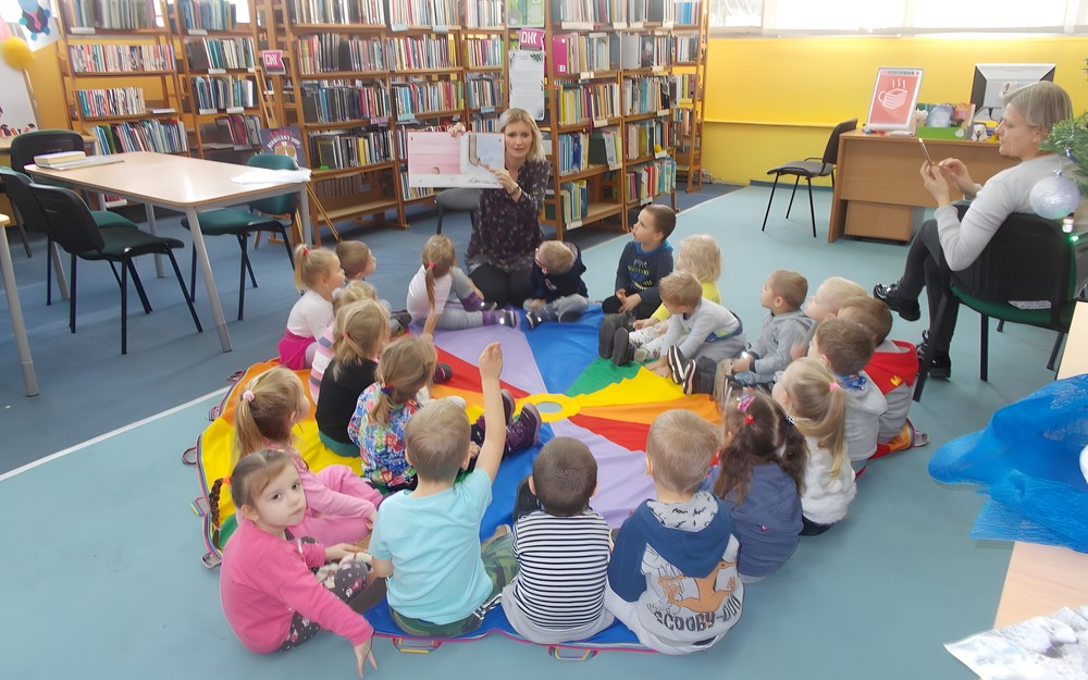 Dzieci siedzą na chuście animacyjnej i słuchają czytnego przez bibliotekarkę opowiadania