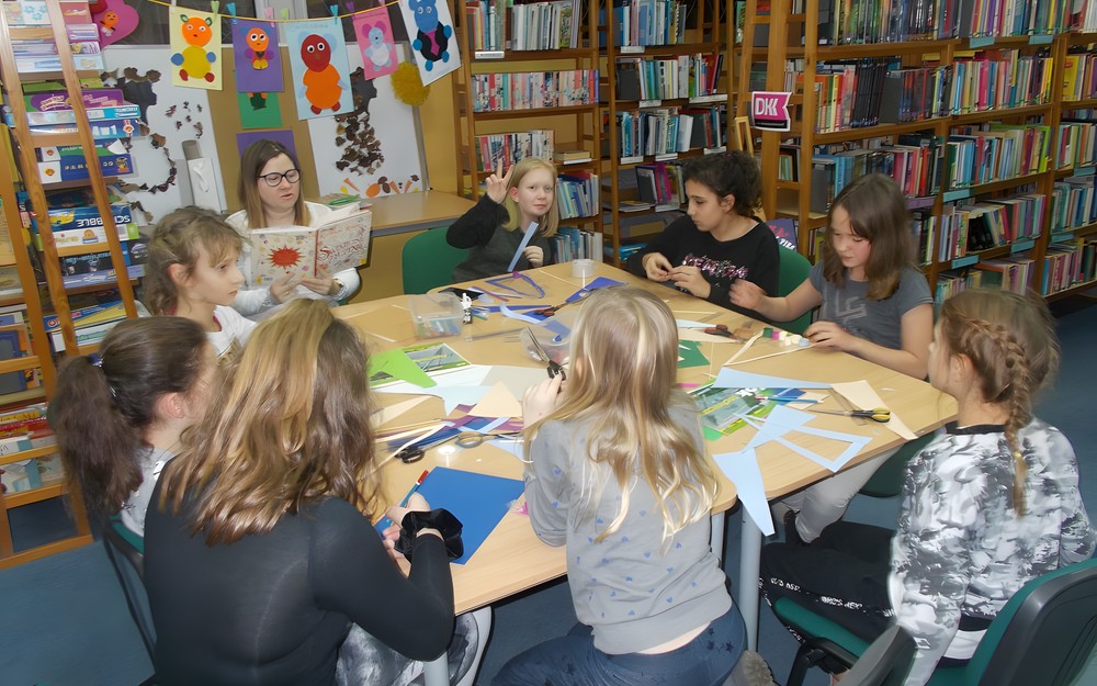 Dzieci siedzą przy stole i wykonują prace plastyczną, podczas gdy bibliotekarka czyta opowieść