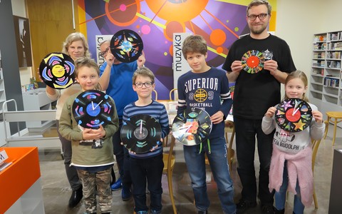 Wspólne zdjęcie. Uczestnicy warsztatów "Czas na muzykę" stoją na tle muralu w Mediatece, każdy trzyma w ręku wykonany przez siebie zegar z płyty winylowej.