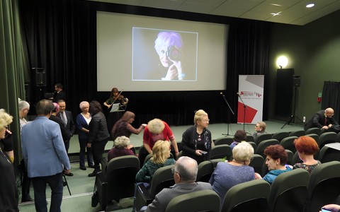 Publiczność gromadząca się na wernisaż wystawy. W tle widać zdjęcie Pani Ewy Maliszewskiej z aparatem w dłoni.