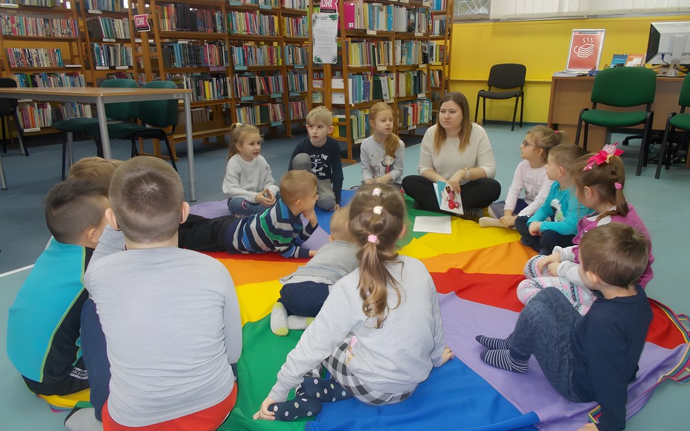 Dzieci siedzą na chuście animacyjnej i słuchają czytnego przez bibliotekarkę opowiadania
