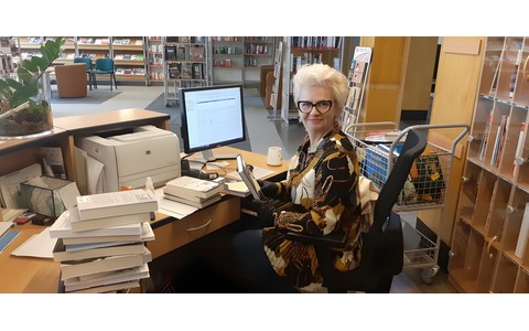 Bibliotekarze nadrabiają zaległości. Bibliotekarka przy biurku pełnym książek.