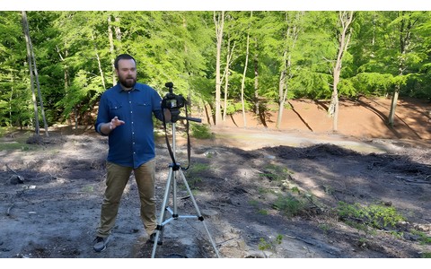 Archeolog Andrzej Kuczkowski stoi w lesie, przed nim na statywie umieszczony jest aparat