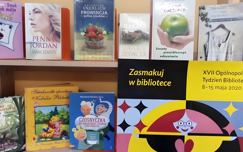 Zdjęcie przedstawia plakat Tygodnia Bibliotek 2020 "Zasmakuj w bibliotece", wraz z książkami o tematyce kulinarnej.ami